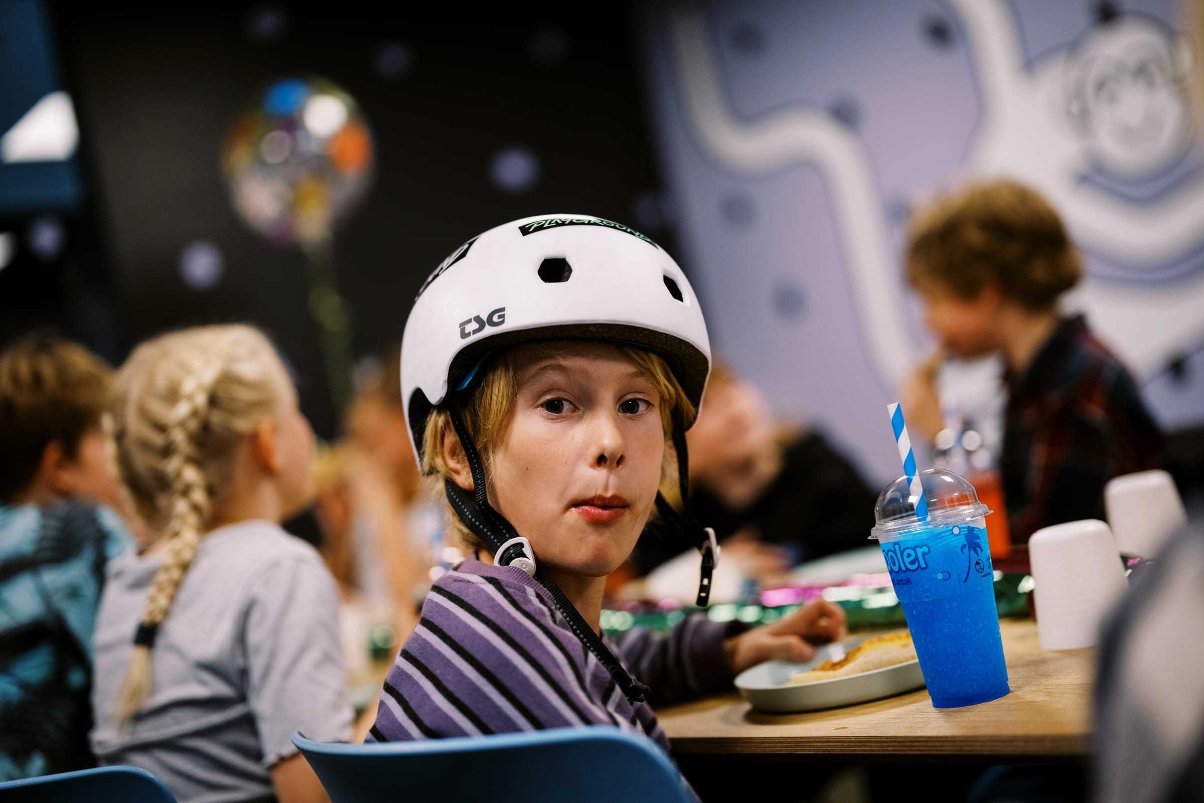 A birthday kid in a helmet with a blue slushie at the table, party balloon behind