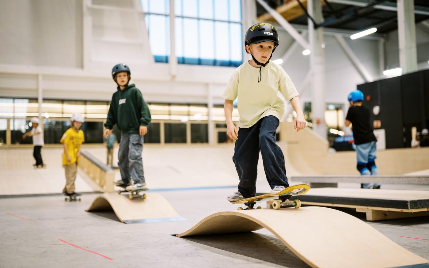 Gutt med hjelm balanserer på skateboard over en trebølge i skatehallen