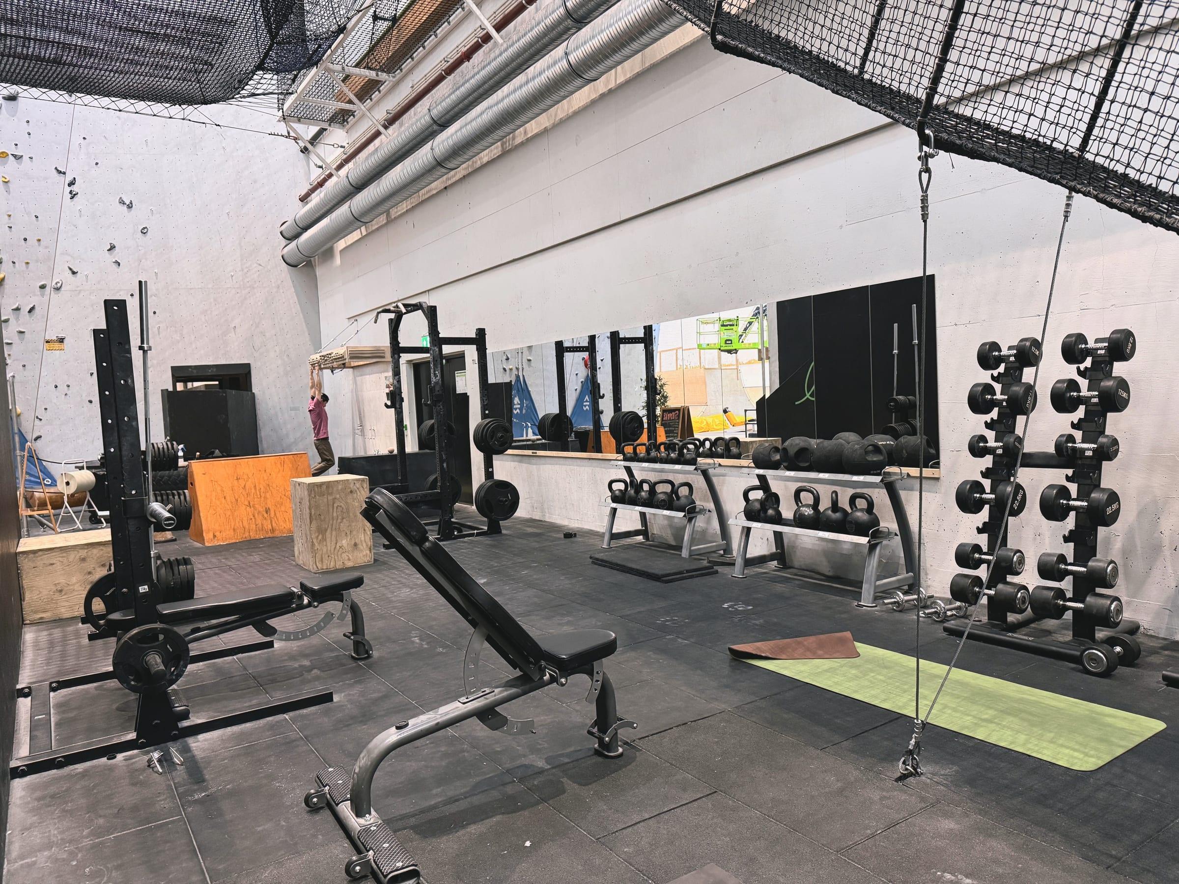 Gymmen på Playground: benker, kettlebells, manualstativ og knebøystativ foran klatreveggen, med en person som henger i et fingerbrett i bakgrunnen