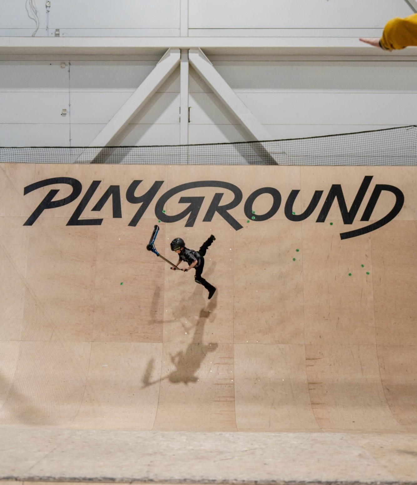 Barn på sparkesykkel mid-air foran PLAYGROUND-logoen på halfpipen